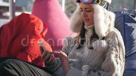 一个女孩用智能手机坐在滑雪场的咖啡馆里。 年轻女子在高山滑雪场放松。视频