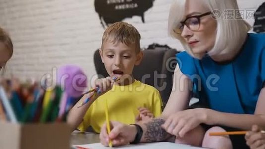 一个穿着黄色t恤，带着彩色两支铅笔的小男孩坐在桌子旁，看着他的老师画画。 绘图视频