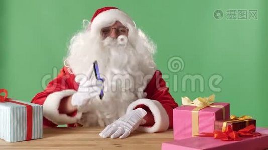 微笑圣诞老人在镜头中展示智能手机，礼物在桌子上，背景中的色度。视频