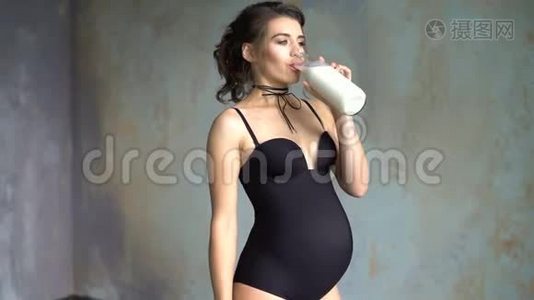 孕妇从杯子里贪婪地喝牛奶。 4K的灰色墙底上美丽的黑发少女视频