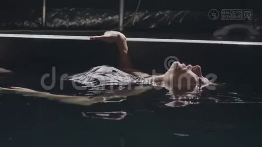 年轻女子躺在黑暗背景下的游泳池里光滑的水面上视频