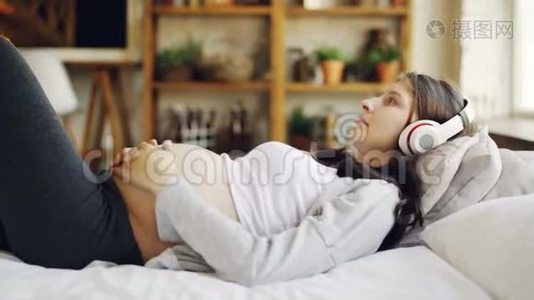 迷人的孕妇正在家里用耳机躺在床上抚摸肚子听音乐。闲暇视频