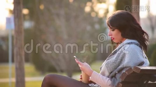 漂亮可爱的黑发女孩坐在城市公园的长凳上，喝热咖啡，里面有蒸汽和写在电话里视频
