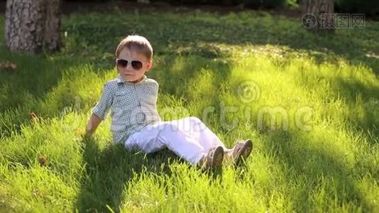 一个戴着太阳镜的可爱小男孩坐在公园的绿草地上。视频