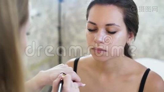 一位年轻的金发碧眼的化妆师用一个遮瑕膏在她脸上画一个年轻的黑发女孩视频