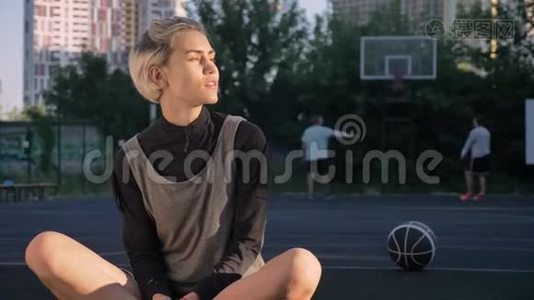 漂亮的金发女人坐在篮球场上，对着镜头微笑，男人在后台打球，女球员视频