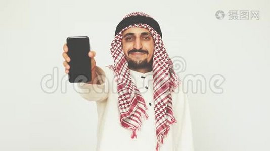 穿着民族服装微笑的阿拉伯人展示了智能手机和广泛的微笑。 一个男人展示一部现代手机。 商业和办公室。 阿拉伯视频