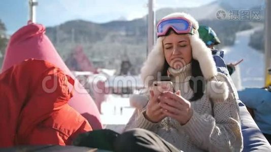在高山滑雪场放松的年轻女子。一个女孩坐在滑雪场的咖啡馆里用智能手机。视频