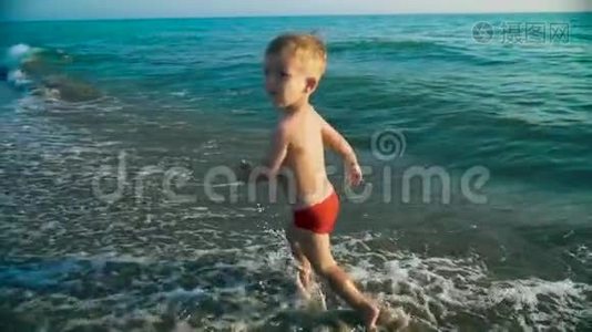 快乐的男孩快乐地跑着跳着，在夕阳下溅在海水里。 慢动作视频