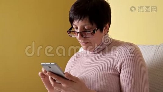 一个戴眼镜的女人在家里抱着一个白色枕头坐在沙发上，在她全新的智能手机上写着信息视频