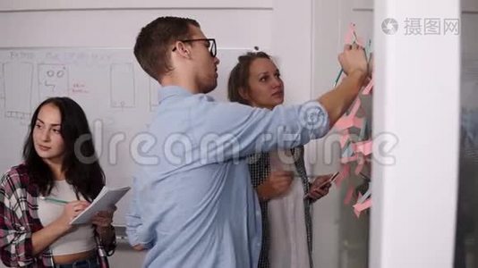 由三个年轻人组成的团队-戴眼镜的年轻人把彩色的纸粘在一起，并在上面做一个笔记。 同事视频