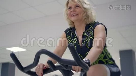 中年金发女人正在骑着固定的自行车做运动。 体育理念。视频
