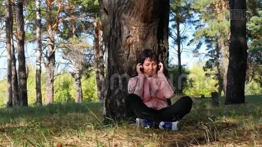 年轻女子坐在公园的一棵树旁，通过耳机听音乐。 音乐、户外娱乐视频