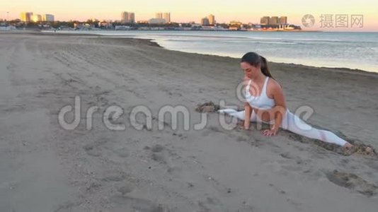 日出时，坐在沙滩上做瑜伽的年轻女子在沙滩上劈叉。视频