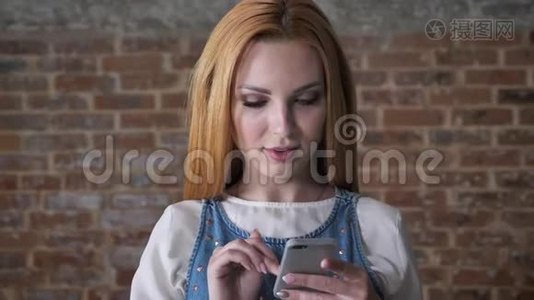 年轻迷人的金发女孩正在智能手机上输入信息，在镜头下观看，砖背景，交流理念视频