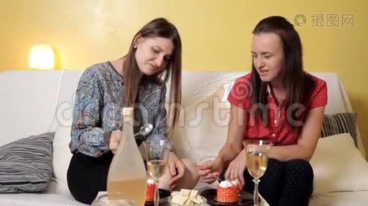 两个年轻女孩坐在家里的沙发上喝着葡萄酒，吃着蛋糕，聊天。 友谊，甜蜜视频
