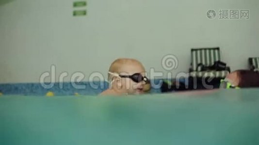 侧面观看，水下拍摄的年轻女子穿着粉红色泳衣，在游泳池里支撑着他的儿子。 如何教学视频