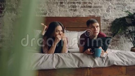年轻人正在看电视，他的女朋友躺在床上拿着遥控器，他们拿着枕头和杯子视频