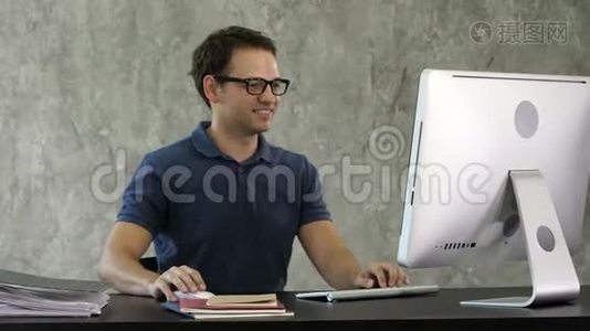微笑的年轻人坐在办公室的办公桌前，在电脑上工作。视频