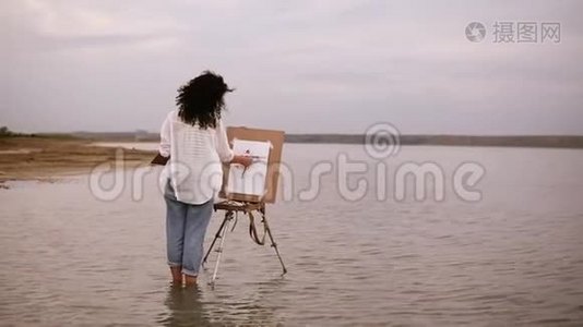 这位穿着蓝色牛仔裤和白色衬衫的女艺术家站在水里画架，直到脚踝，为她的未来工作视频