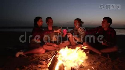 一群人晚上在海滩上的篝火旁消磨时间。 喝酒，干杯。 年轻人抱着视频