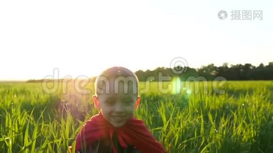 一个穿着超级英雄服装，穿着红色斗篷的快乐孩子坐在一片绿色的草坪上，映衬着夕阳，然后升起视频