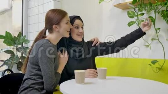 两个年轻美丽的穆斯林妇女坐在咖啡馆里，其中一个戴着头巾，自拍，看着电话，微笑着，视频