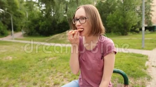 戴眼镜的滑稽时髦女孩坐在长凳上吃一片比萨饼。视频