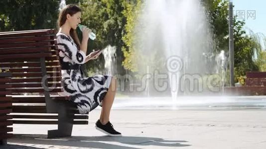年轻的女商人坐在长凳上喝水，在平板电脑上工作视频