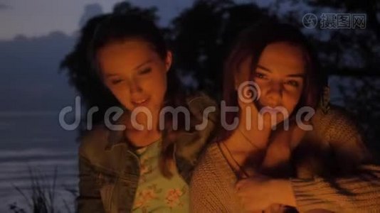 两个白种人女孩晚上坐在大自然的炉火旁，看着摄像机，微笑着，思考着视频