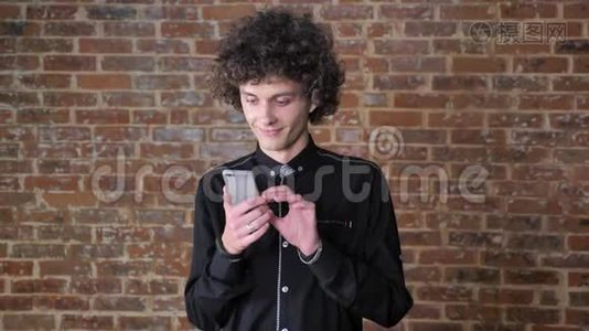 一位留着卷发的年轻帅哥用他的手机站在砖墙附近视频