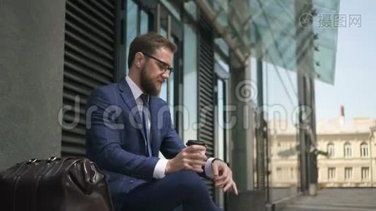 大胡子的商人看着手表，坐在城市街道上喝着咖啡。视频