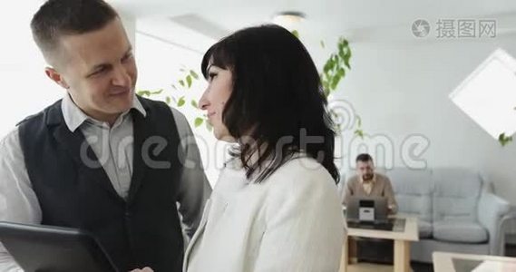 一个穿西装的年轻人和一个中年妇女站在办公室大厅里，拿着一块平板电脑，讨论交易的细节。视频
