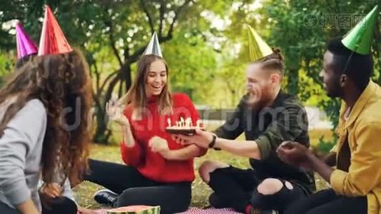 戴着派对帽的漂亮女孩在公园野餐时和朋友们一起庆祝生日许愿，在蛋糕上吹蜡烛视频