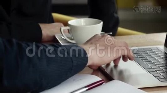 商人的双手，在咖啡馆里坐在木桌旁，可以看到一个在笔记本电脑上工作的人。 讨论视频