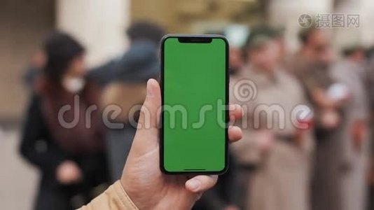 一名男子手持一部移动电话的特写镜头，街道上有一个垂直的绿色屏幕。大屏幕。军事视频