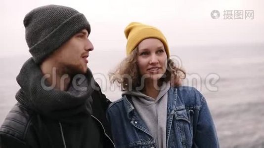 一对年轻的幸福夫妇站在一起，在海边拥抱。 穿暖和的衣服，戴冷帽的年轻人视频