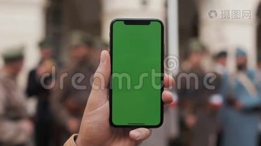 一名男子手持一部移动电话的特写镜头，街道上有一个垂直的绿色屏幕。大屏幕。军事视频