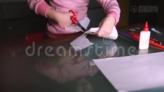 一张可爱的学龄前女孩的特写照片，她穿着粉色毛衣，用剪刀从纸上剪下形状，反射在玻璃桌上。视频