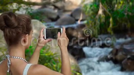 年轻女子在丛林森林中的山瀑布中用手机做照片景观。 女游客视频