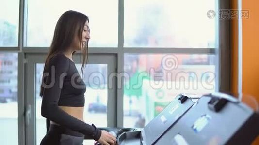 一位年轻的黑发女子在健身房的跑步机上站在窗口背景下视频