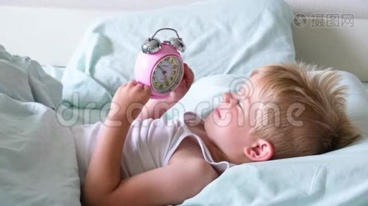 一个金发小男孩躺在床上微笑，一大早就玩闹钟。 那男孩听着钟响视频