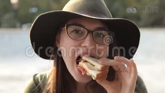 一个戴着眼镜和帽子的年轻女孩正在尝试一个美味的三明治。 食物视频