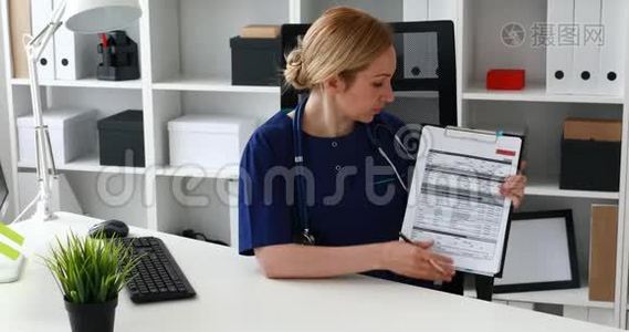 医生坐在办公椅上，在文件中显示笔记，看着相机。视频