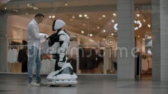 一个穿衬衫的男人和一个白色机器人交流，他问问题，用手指按屏幕。视频