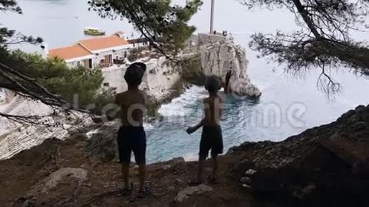 两个男孩站在高高的岩石上欣赏大海的后景。 两个哥哥站在高处的剪影视频