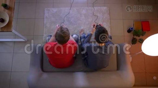 两个穿着睡衣的年轻人用操纵杆坐在客厅的沙发上玩游戏。视频