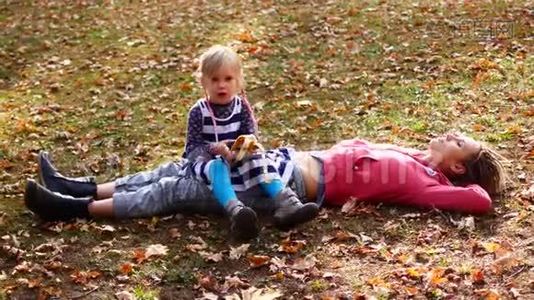 美丽的妈妈和可爱的孩子躺在秋天公园的草地上。 女儿坐在妈妈身上吃香蕉，妈妈望着天空视频