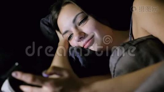 女孩晚上躺在卧室的床上上网，看照片，聊天和微笑。视频