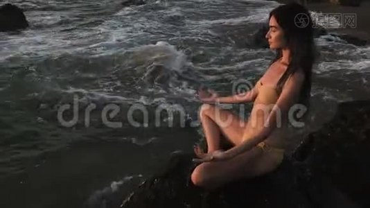 女孩坐在海边的岩石上，沉思着莲花瑜伽的姿势，全神贯注。视频